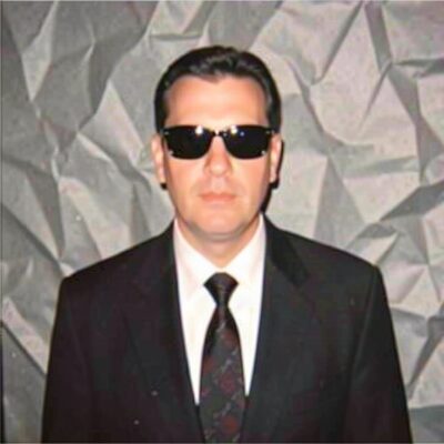 A man in a suit and sunglasses in front of a grey backdrop.